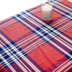 May include: A red, blue, and white plaid tablecloth with a lit candle in a glass holder. The tablecloth features a traditional plaid design with intersecting lines of red, blue, and white. A candle burns in a clear glass container.
