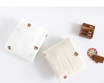 Könnte beinhalten: Zwei weiße und cremefarbene Musselin-Babydecken mit einem braunen Bären-Druck. Die Decken sind gefaltet und auf einer weißen Oberfläche platziert.