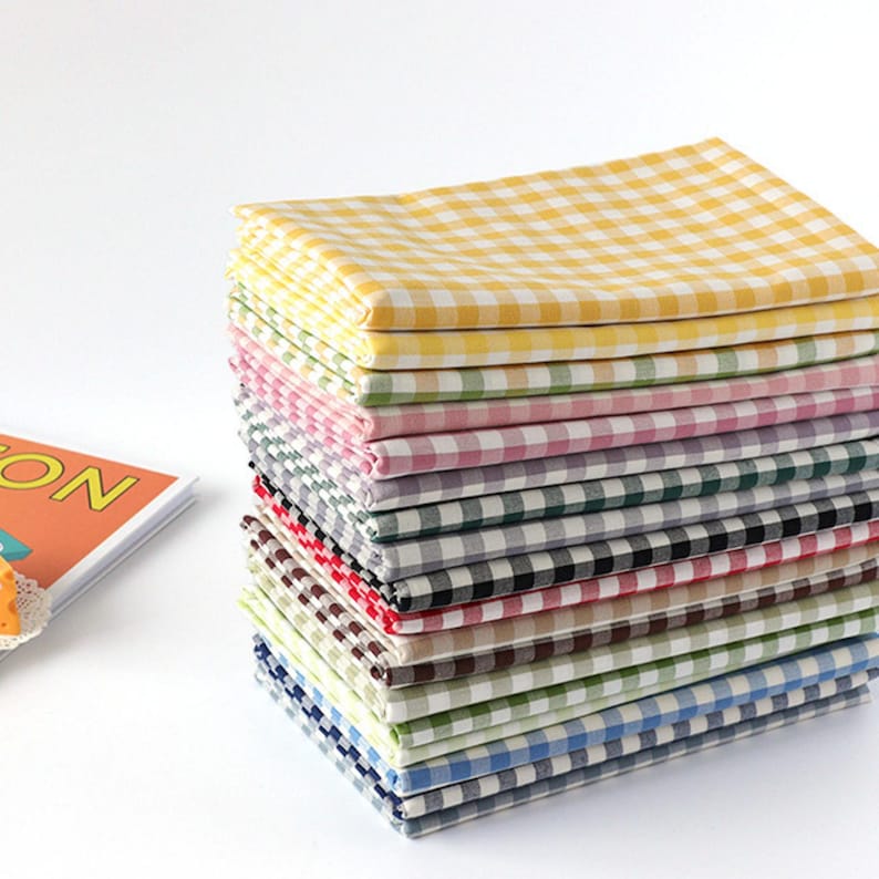 May include: A stack of colorful checkered fabric napkins in various colors, including yellow, green, pink, gray, black, brown, and blue. The napkins are neatly folded and stacked, creating a visually appealing display. The fabric has a classic gingham pattern.