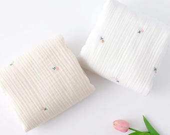 Könnte beinhalten: Zwei gefaltete quadratische Musselintücher, eines in Creme und eines in Weiß, beide mit einer dezenten vertikalen Knitterstruktur und einem wiederkehrenden Muster aus kleinen gestickten rosa Tulpen mit grünen Stielen, ergänzt durch eine einzelne rosa Tulpenblüte.