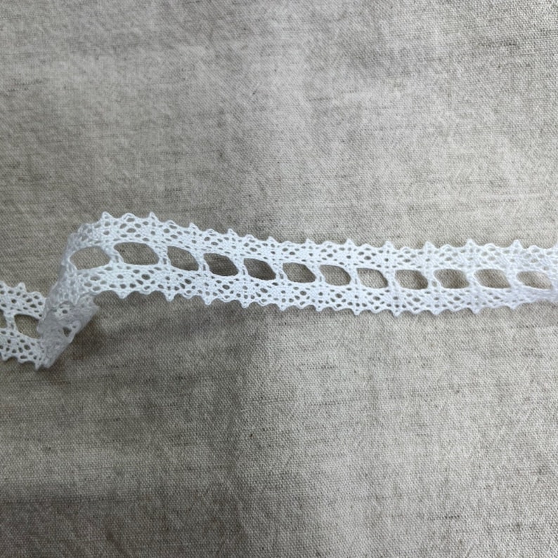 May include: White lace trim featuring a delicate diamond pattern. The trim is made from a soft, lightweight material and has a slightly scalloped edge.