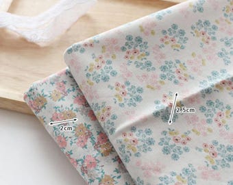 May include: Two folded pieces of white fabric featuring a repeating floral pattern in shades of pink, yellow, and teal. One section shows a 2cm measurement, while another section indicates a 2.5cm measurement with black arrows.