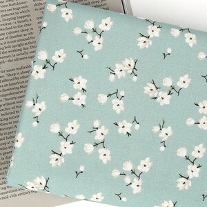 May include: A light sage green fabric featuring a repeating pattern of small white blossoms with dark brown stems and branches. The floral print is evenly distributed across the surface, showcasing delicate petals and buds in a consistent arrangement.
