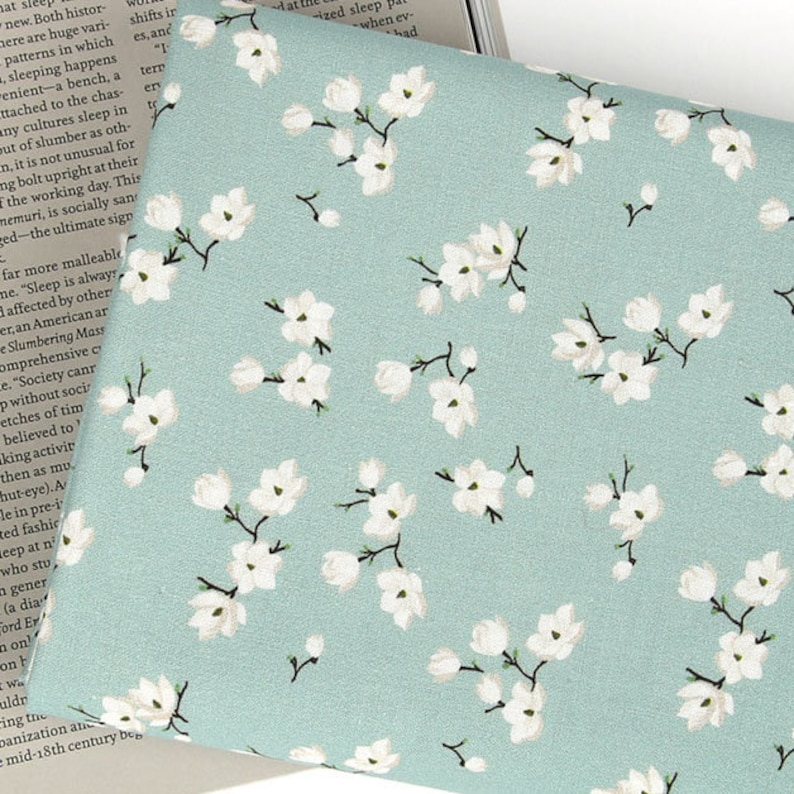 May include: A light sage green fabric featuring a repeating pattern of small white blossoms with dark brown stems and branches. The floral print is evenly distributed across the surface, showcasing delicate petals and buds in a consistent arrangement.