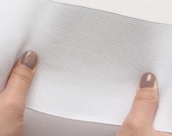 May include: Close-up of a white elastic band, held between two fingers. The elastic appears to be thick and sturdy, with a woven texture. The fingers have light-coloured nail polish.