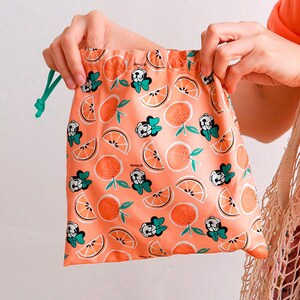Puede incluir: Una bolsa de cord&oacute;n rosa con un patr&oacute;n repetido de rodajas de naranja y un personaje de dibujos animados con un lazo. La bolsa tiene un cord&oacute;n de color verde.