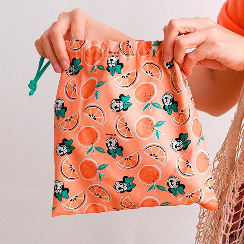 Puede incluir: Una bolsa de cord&oacute;n rosa con un patr&oacute;n repetido de rodajas de naranja y un personaje de dibujos animados con un lazo. La bolsa tiene un cord&oacute;n de color verde.