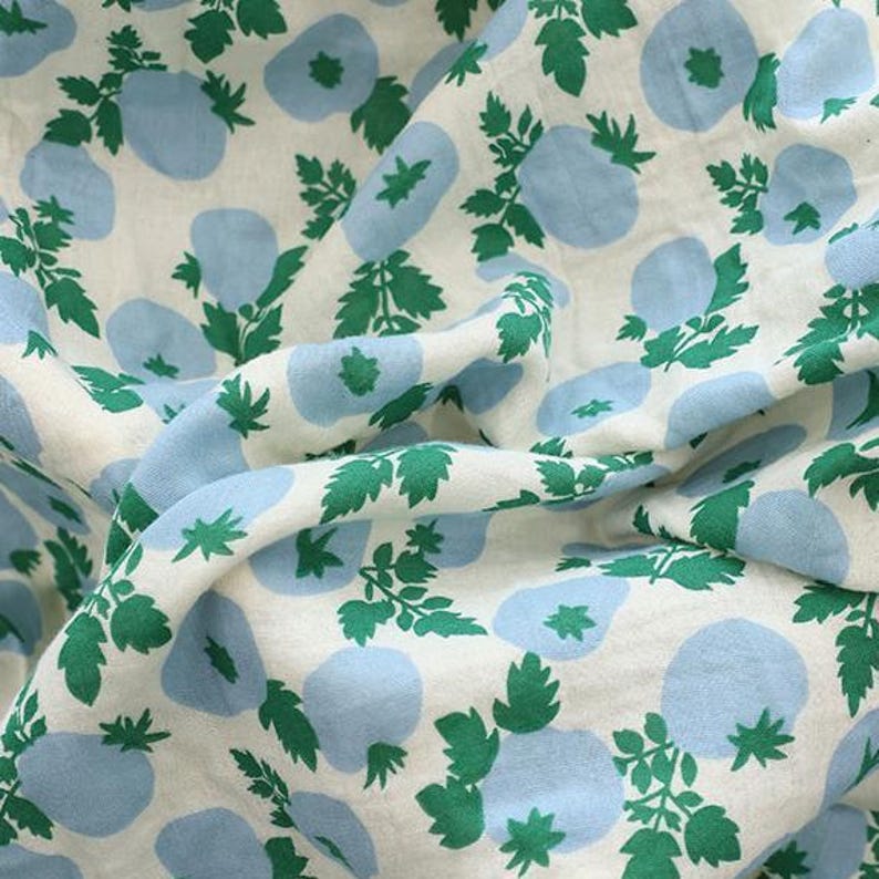 May include: A piece of off-white fabric featuring a repeating pattern of light blue rounded shapes and vibrant green leafy stems. The textile is draped to show folds and texture, highlighting the contrast between the blue and green print.