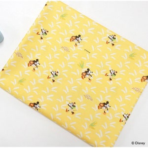 Puede incluir: Tela amarilla con un patrón repetido de Mickey Mouse y Minnie Mouse caminando por un campo de trigo. El texto "© Disney" aparece en la esquina superior derecha de la tela.
