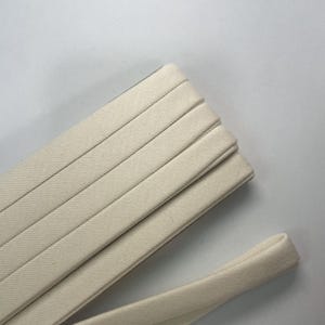 Pu&ograve; includere: Un rotolo di nastro in twill di cotone color bianco sporco piegato in strati ordinati e sovrapposti. Il tessuto presenta un distinto motivo a trama diagonale e una consistenza liscia e opaca. Un'estremit&agrave; del nastro si estende verso l'esterno.