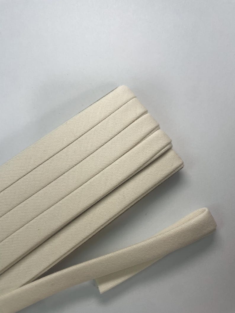 Pu&ograve; includere: Un rotolo di nastro in twill di cotone color bianco sporco piegato in strati ordinati e sovrapposti. Il tessuto presenta un distinto motivo a trama diagonale e una consistenza liscia e opaca. Un'estremit&agrave; del nastro si estende verso l'esterno.