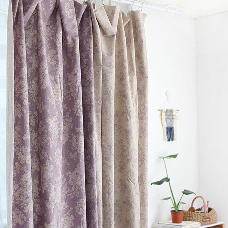 K&ouml;nnte beinhalten: Bodenlange Vorh&auml;nge mit einem sich wiederholenden Blumenmuster in gedeckten Violett- und Beiget&ouml;nen. Der Stoff h&auml;ngt an einer wei&szlig;en Stange mit kreisf&ouml;rmigen Ringen und bildet vertikale Falten. Das Design zeigt einen durchgehenden botanischen Druck.