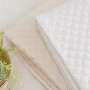 May include: Two folded quilted blankets stacked on top of each other. The top blanket is bright white, and the bottom blanket is a soft beige colour. Both blankets feature a consistent diamond-shaped stitched pattern across their entire surface.