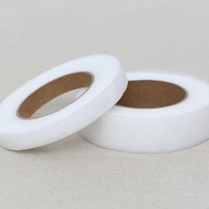 May include: Two rolls of white, non-woven interfacing fabric. The rolls are circular with a brown cardboard core. One roll is partially unrolled, revealing the core. The fabric has a soft, slightly textured appearance.