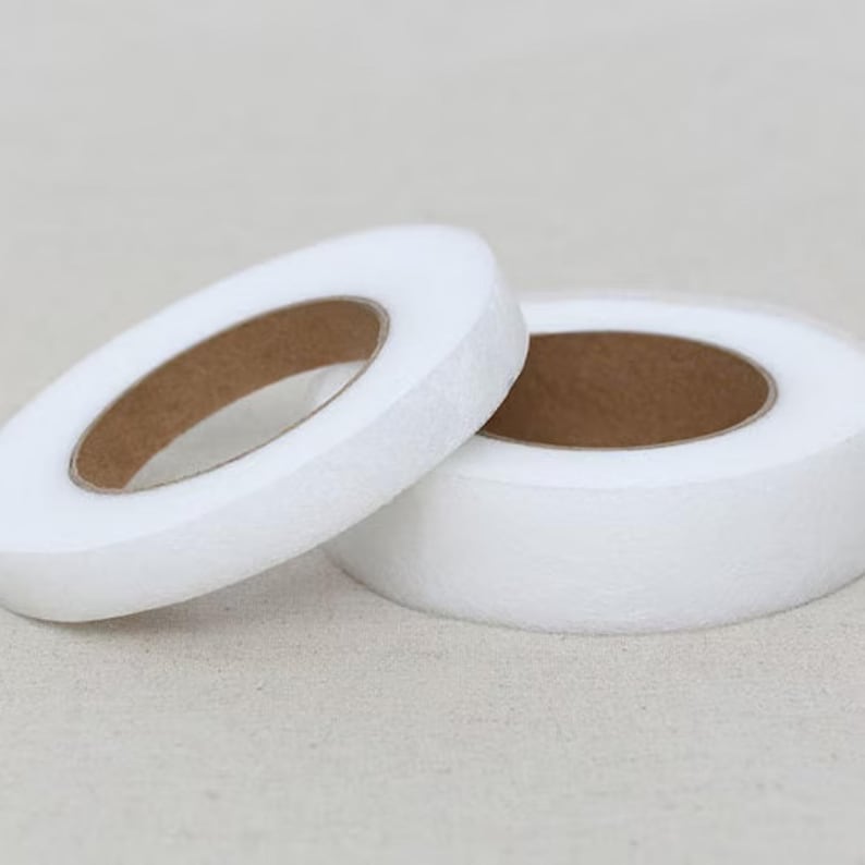 May include: Two rolls of white, non-woven interfacing fabric. The rolls are circular with a brown cardboard core. One roll is partially unrolled, revealing the core. The fabric has a soft, slightly textured appearance.