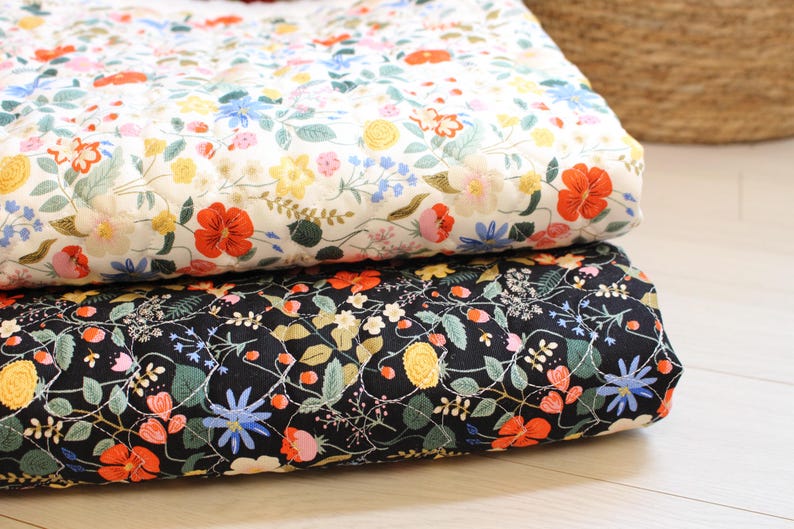 Pu&ograve; includere: Due coperte trapuntate piegate con un motivo floreale. La coperta superiore ha uno sfondo bianco con fiori rossi, gialli e blu. La coperta inferiore ha uno sfondo nero con la stessa stampa floreale, che include foglie e steli di colore verde.