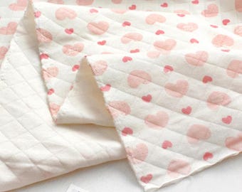 May include: A soft, off-white quilted fabric featuring a repeating pattern of small, light pink hearts. The material has a diamond-stitched texture throughout, creating a subtle grid effect across the surface of the textile.