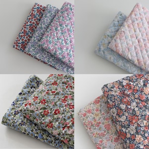 May include: A collection of folded fabric swatches featuring various floral patterns. The textiles display intricate botanical prints in shades of red, blue, pink, green, and white, with some pieces showcasing a quilted diamond-stitched texture.