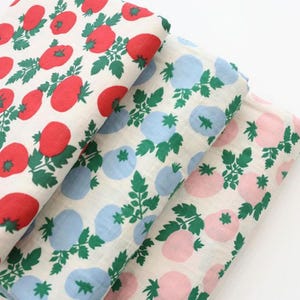 May include: Three folded pieces of fabric featuring a repeating pattern of tomatoes and green leaves. The first piece has red tomatoes on a cream background, the second has light blue tomatoes, and the third has light pink tomatoes on cream.