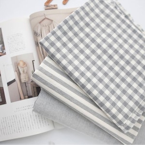 Könnte beinhalten: Ein Stapel Stoffmuster in verschiedenen Mustern. Der obere Stoff hat ein grau-weißes Vichy-Karo-Muster. Darunter befindet sich ein Stoff mit grau-weißen Streifen und unten ein einfarbiger grauer Stoff. Im Hintergrund ist ein offenes Magazin zu sehen.