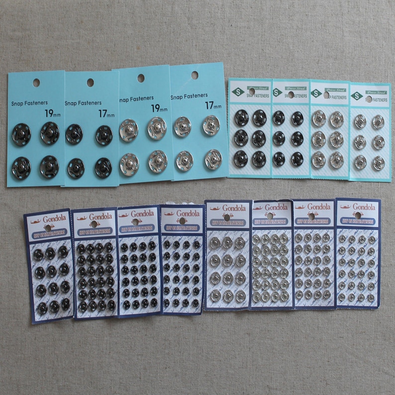 May include: Assortment of silver and black snap fasteners in various sizes, packaged on cards labelled "Snap Fasteners" and "Gondola". The cards show the size of the fasteners in millimetres, such as 17mm and 19mm.