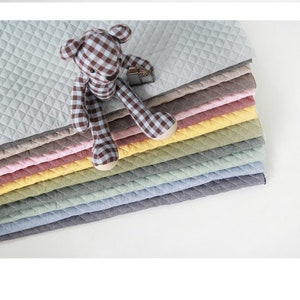 May include: A stack of quilted fabric mats in various pastel colours including light grey, beige, pink, yellow, sage green, light blue, and dark grey, topped with a brown and white gingham patterned teddy bear with solid beige paws.
