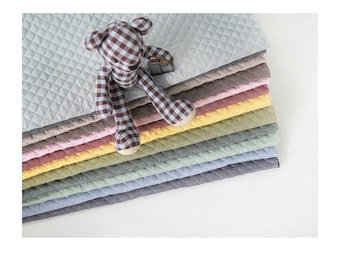 Puede incluir: Una pila de alfombrillas de tela acolchada en varios colores pastel, incluyendo gris claro, beige, rosa, amarillo, verde salvia, azul claro y gris oscuro, coronada por un oso de peluche con patrón de cuadros marrón y blanco.