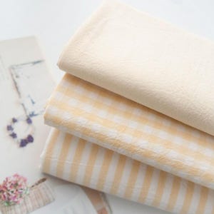 Peut inclure: Trois morceaux de tissu dans des motifs différents : un tissu uni jaune clair, un tissu vichy jaune et blanc, et un tissu rayé jaune et blanc.