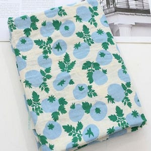 May include: A folded piece of fabric featuring a repeating pattern of light blue circular shapes and green leafy stems on an off-white background. The textile displays a consistent botanical print across the entire visible surface area.