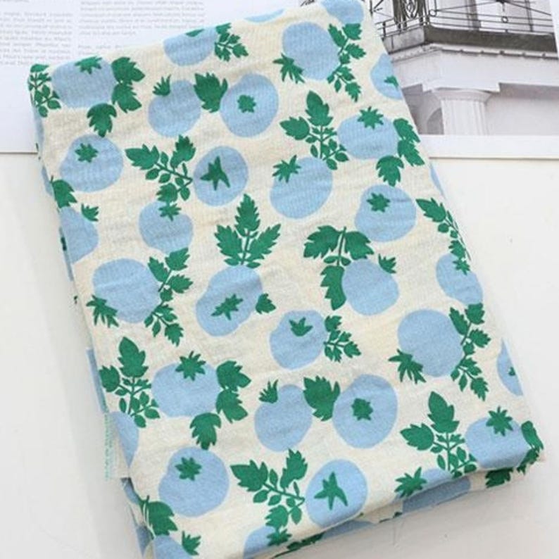 May include: A folded piece of fabric featuring a repeating pattern of light blue circular shapes and green leafy stems on an off-white background. The textile displays a consistent botanical print across the entire visible surface area.