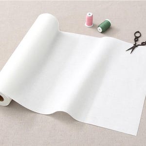 May include: A roll of white fabric with a brown cardboard core. The fabric is unrolled and folded in half. There are two spools of thread, one pink and one green, and a pair of black scissors on the fabric.