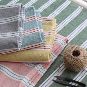 May include: Four different striped fabric swatches in grey, blue, yellow, and pink, stacked on top of each other. The fabric swatches are on a green and white striped surface. A ball of twine is in the bottom right corner of the image.