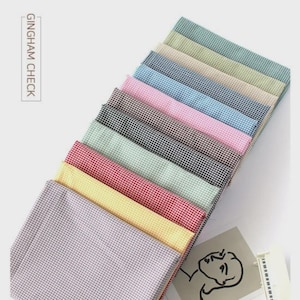 Può includere: Una pila di tessuto a quadretti vichy in vari colori, tra cui rosa, blu, nero e rosso. Il tessuto è disposto in diagonale, mostrando le diverse opzioni di colore. Il testo "GINGHAM CHECK" è visibile sul lato sinistro.