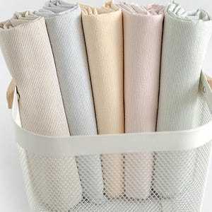 Pode incluir: Cinco rolos de tecido canelado em tons pastel bege, azul claro, amarelo pálido, rosa suave e verde menta estão organizados dentro de uma cesta de malha metálica branca com alças de madeira, exibindo uma textura vertical e forma cilíndrica.
