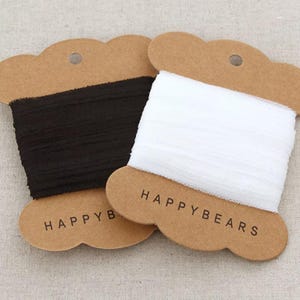 May include: Two spools of ribbon, one black and one white, are displayed on brown cardboard tags. The tags are shaped like a cloud with the words "HAPPYBEARS" printed on them. The ribbon is tightly wound.