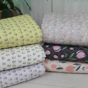 May include: A stack of quilted blankets in various floral patterns. The blankets feature a yellow, white, and lavender base with small flower designs. Other blankets have a gray base with pink tulip designs and a white base with pink tulip designs.