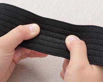 May include: Close-up of a black elastic band held between two hands. The band has a ribbed texture and appears to be made of a durable material. The background is a neutral, light colour.