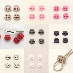 May include: Assortment of plastic toggle clasps in various colors, including black, white, red, pink, green, and beige. The clasps have two holes and are used for securing cords, straps, or other materials.