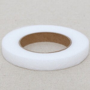 May include: A roll of white, non-woven interfacing fabric. The interfacing is a ring shape with a brown cardboard core. The fabric is thick and appears to be soft. The roll is sitting on a light beige surface.