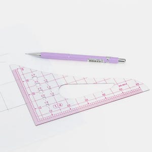 Peut inclure: Un triangle de dessin en plastique transparent avec des marquages roses et un crayon mécanique violet. Le triangle comporte des mesures en centimètres et une découpe incurvée. Le crayon repose sur une surface blanche.