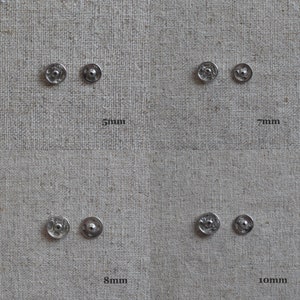 May include: Four silver metal snap fasteners in different sizes: 5mm, 7mm, 8mm, and 10mm. The fasteners are displayed on a beige fabric background.