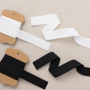 May include: Two spools of flat elastic ribbon, one white and one black, each wrapped around a brown cardboard card. The elastic bands are smooth, opaque, and extend from the spools in loose, flowing folds across the frame.