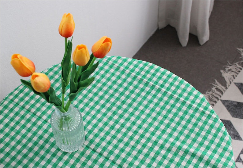 K&ouml;nnte beinhalten: Eine gr&uuml;n-wei&szlig;e karierte Tischdecke mit einem Strau&szlig; orangefarbener Tulpen in einer klaren Glasvase.