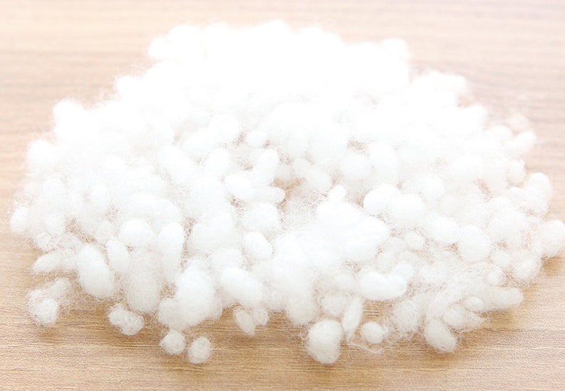 May include: A pile of fluffy, white polyester fiberfill, ideal for stuffing pillows, cushions, or plush toys. The soft, cloud-like material is shown against a light wood grain surface.
