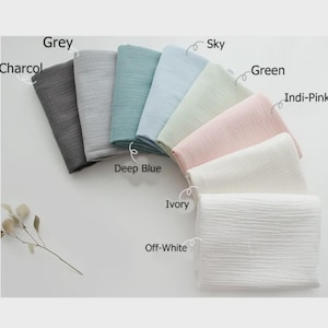 May include: A collection of folded muslin fabric swatches in various colors labeled as Charcoal, Grey, Deep Blue, Sky, Green, Indi-Pink, Ivory, and Off-White. Each piece of fabric features a distinct crinkled texture and is arranged in a fan-like pattern.