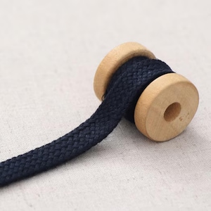 May include: A spool of navy blue braided rope with wooden ends. The rope is tightly wound between two light-colored wooden discs. The rope extends from the spool, showcasing its texture and color. The background is a neutral, off-white fabric.