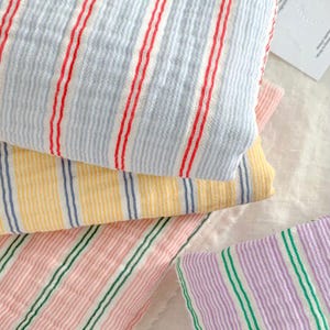 May include: A collection of folded fabric swatches featuring a crinkled texture and vertical stripes. The patterns include light blue with red stripes, yellow with blue stripes, pink with green stripes, and lavender with green stripes.