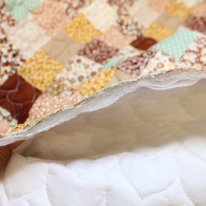 Pu&ograve; includere: Una trapunta patchwork caratterizzata da una variet&agrave; di piccoli quadrati di tessuto in tonalit&agrave; marrone, beige, giallo e verde chiaro. Il retro della trapunta rivela una fodera in tessuto bianco con un motivo a cuori trapuntati e strati di imbottitura.
