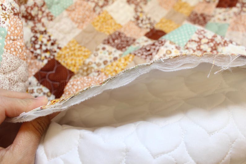 Pu&ograve; includere: Una trapunta patchwork caratterizzata da una variet&agrave; di piccoli quadrati di tessuto in tonalit&agrave; marrone, beige, giallo e verde chiaro. Il retro della trapunta rivela una fodera in tessuto bianco con un motivo a cuori trapuntati e strati di imbottitura.