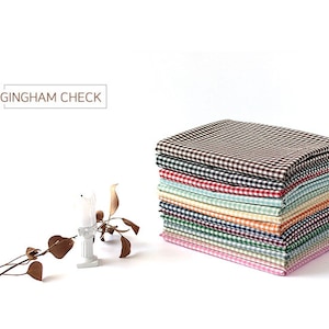 Può includere: Una pila di 10 quadrati di tessuto a quadri vichy di diversi colori. Il tessuto è marrone, rosso, verde, blu, giallo e rosa. Il tessuto è piegato ordinatamente e impilato uno sopra l'altro. Vichy.